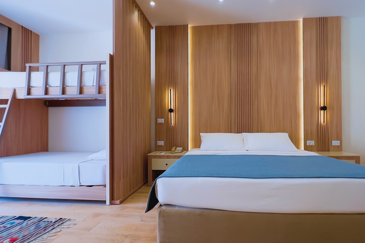 Modern room with wood floors, double bed with blue bedding, bunk beds, wall-mounted TV, and pink accent wall featuring a white palm tree design. Bright and cozy.