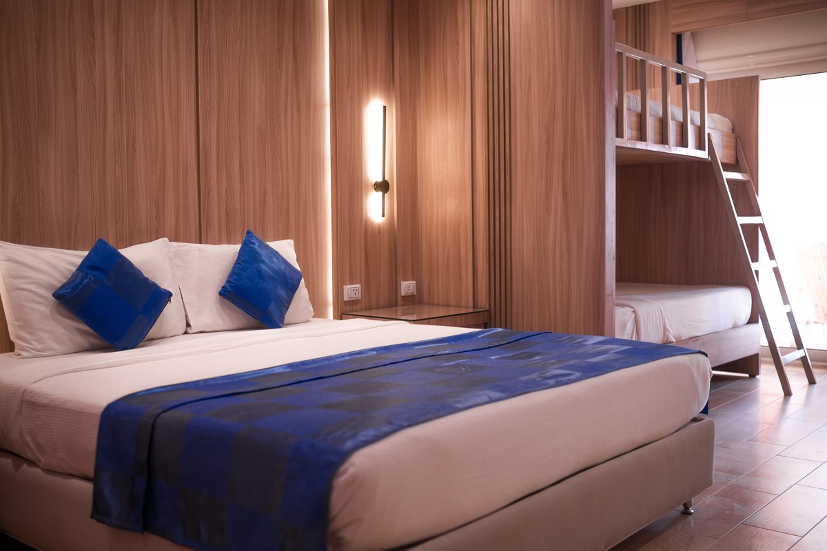 Modern room with wood floors, double bed with blue bedding, bunk beds, wall-mounted TV, and pink accent wall featuring a white palm tree design. Bright and cozy.