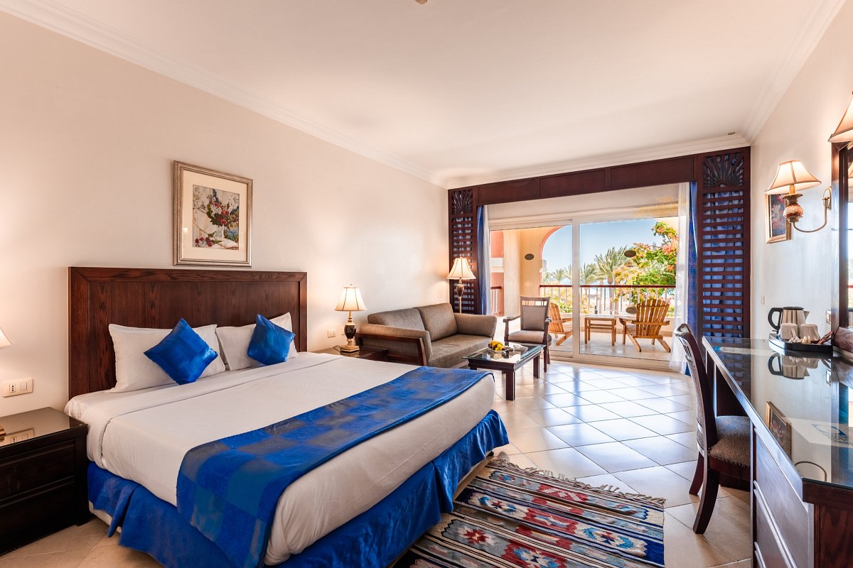 Elegant room with a large bed adorned with blue accents, a cozy sofa, a desk with a chair, and a spacious balcony overlooking greenery. Warm lighting and tasteful decor create a welcoming atmosphere.