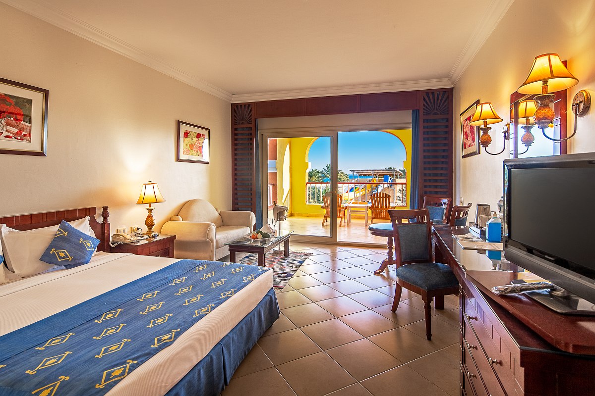 Spacious room with a large bed dressed in blue, a comfortable sofa, tiled floor, desk area, and balcony with garden view. Warm lighting and tasteful decor create a relaxing atmosphere.