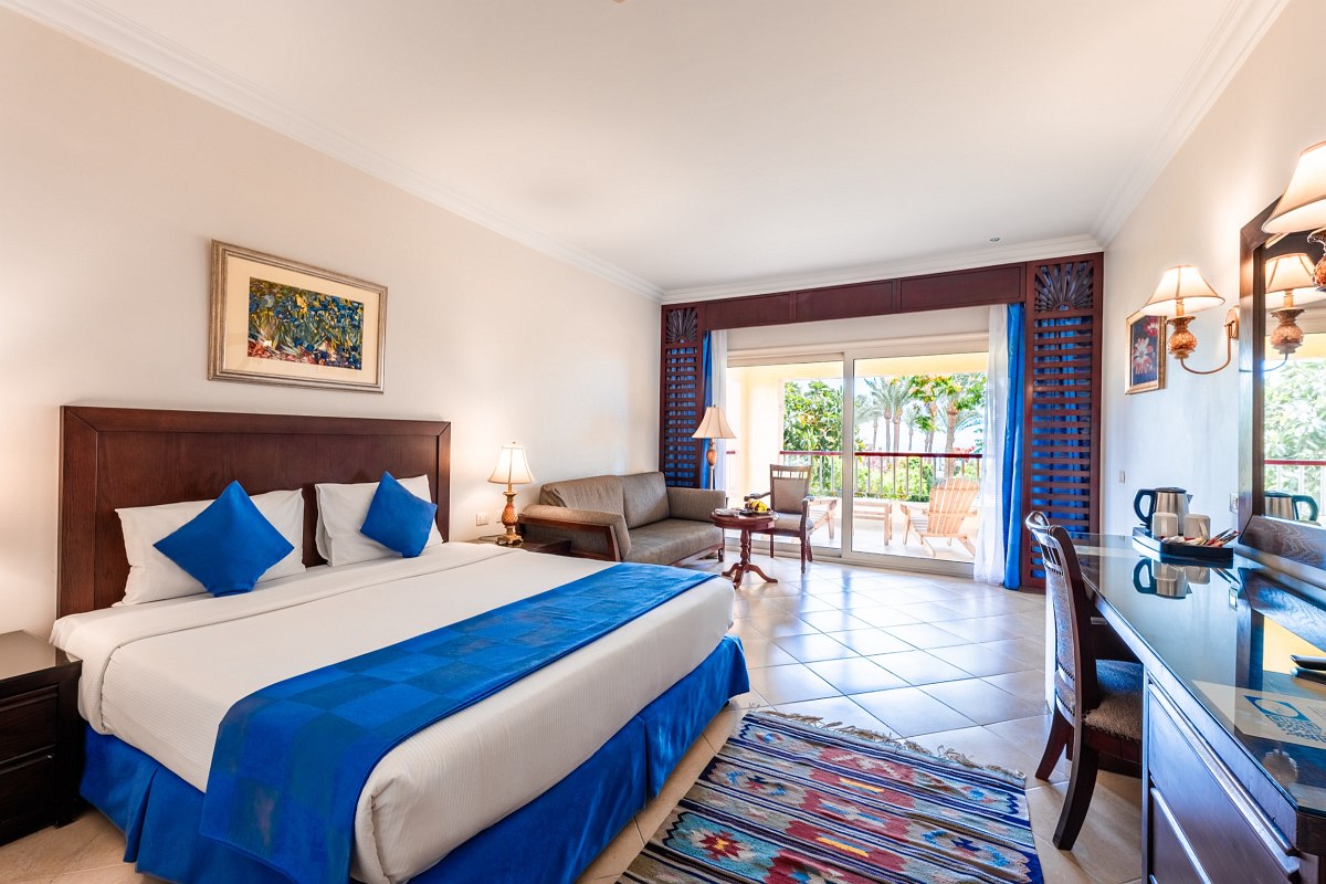 Spacious room with a large bed dressed in blue, a comfortable sofa, tiled floor, desk area, and balcony with garden view. Warm lighting and tasteful decor create a relaxing atmosphere.