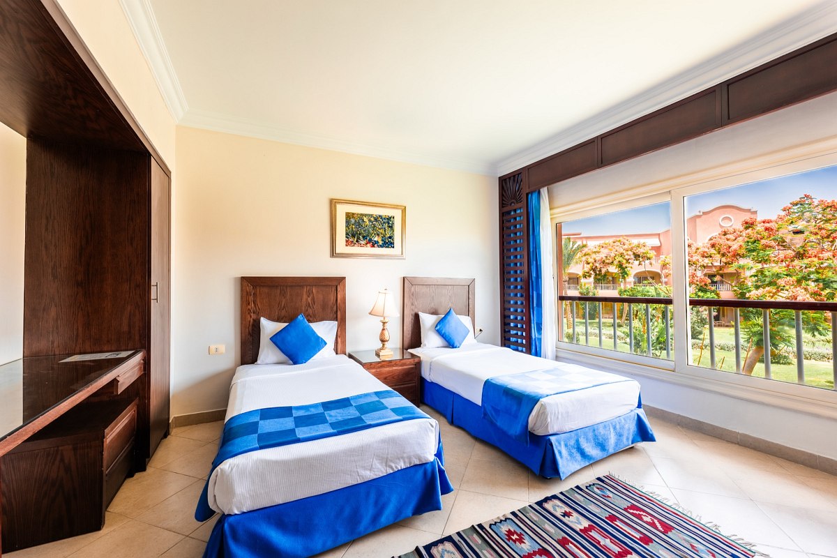 Cozy room with two single beds, blue bedding, a bedside table with a lamp, tiled floor, and a large window offering a garden view. Warm, inviting decor with natural light.