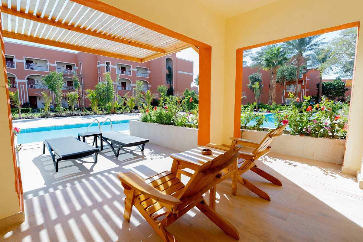 Cozy room with a large bed covered in blue bedding, a comfortable sofa, wooden desk and chair, and a balcony with outdoor seating and a view of greenery. Warm lighting adds to the inviting atmosphere.