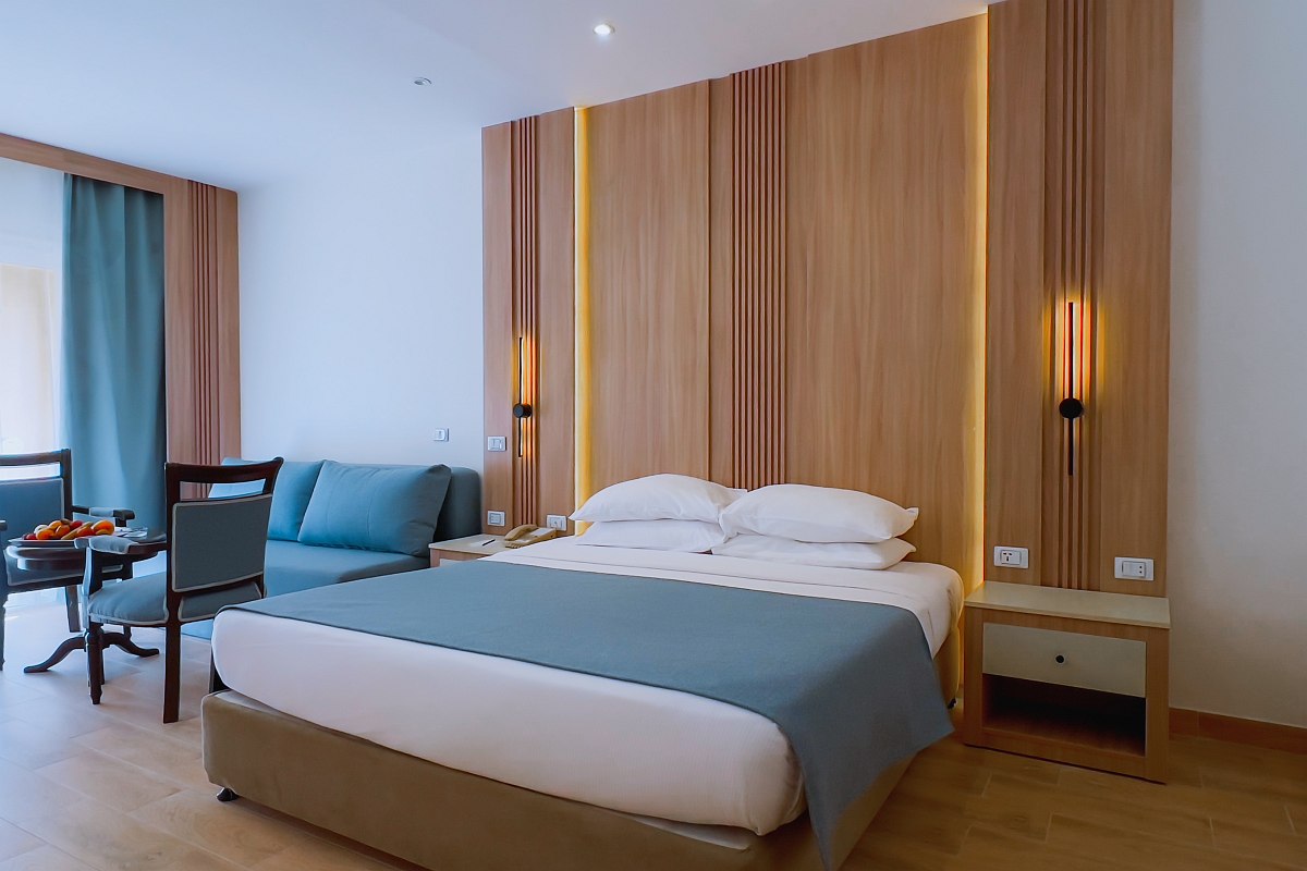 Cozy room with a large bed covered in blue bedding, a comfortable sofa, wooden desk and chair, and a balcony with outdoor seating and a view of greenery. Warm lighting adds to the inviting atmosphere.