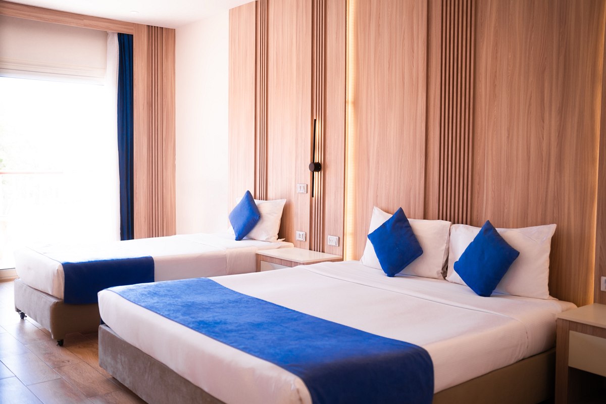 Bright room with two beds, blue accents on pillows and blankets, light wood paneling behind the beds, and a large window letting in natural light. Clean and modern design.