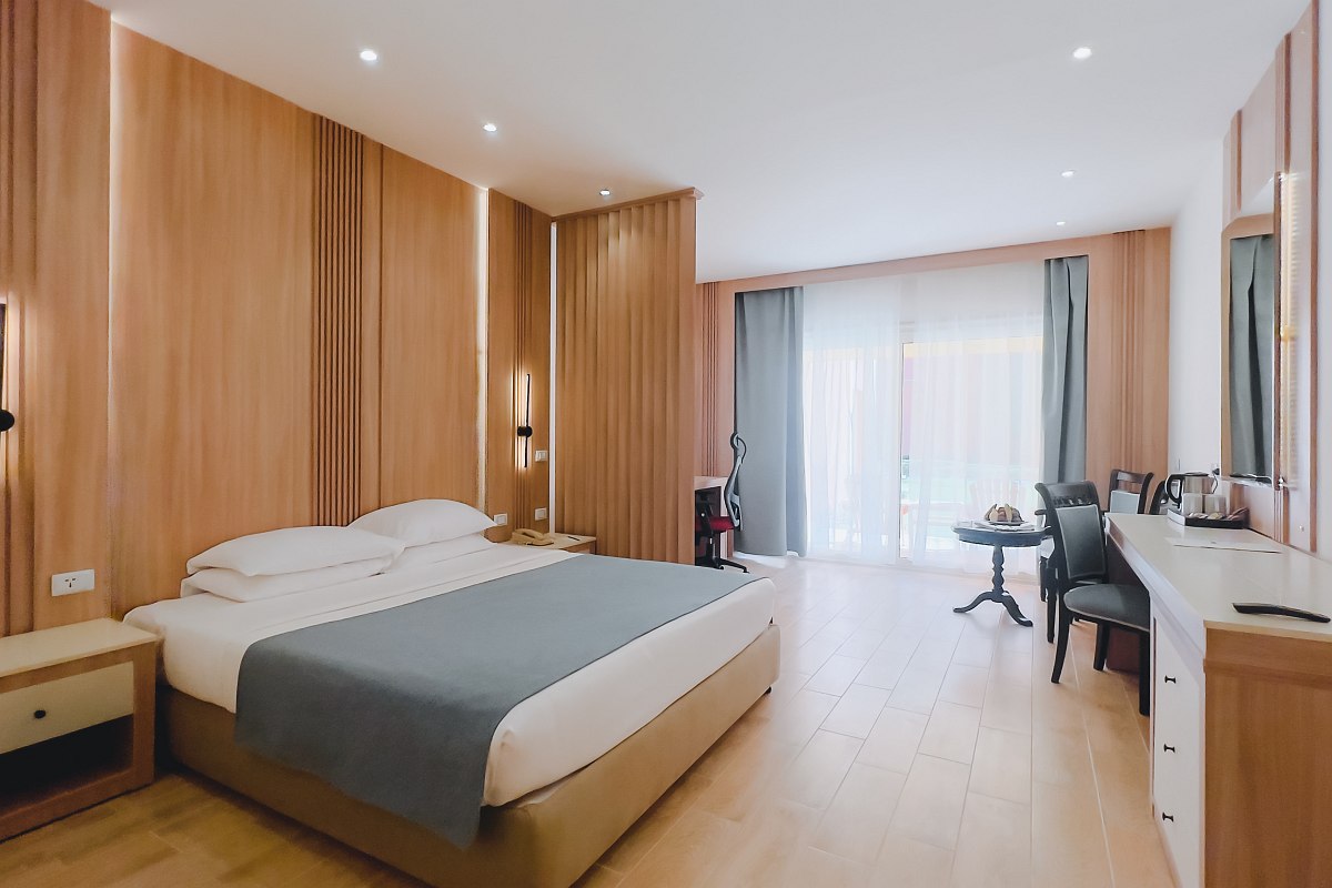 Spacious, modern room with a large bed, wooden panel walls, light wood flooring, and a seating area near a balcony door. Soft lighting and neutral tones create a calm atmosphere.