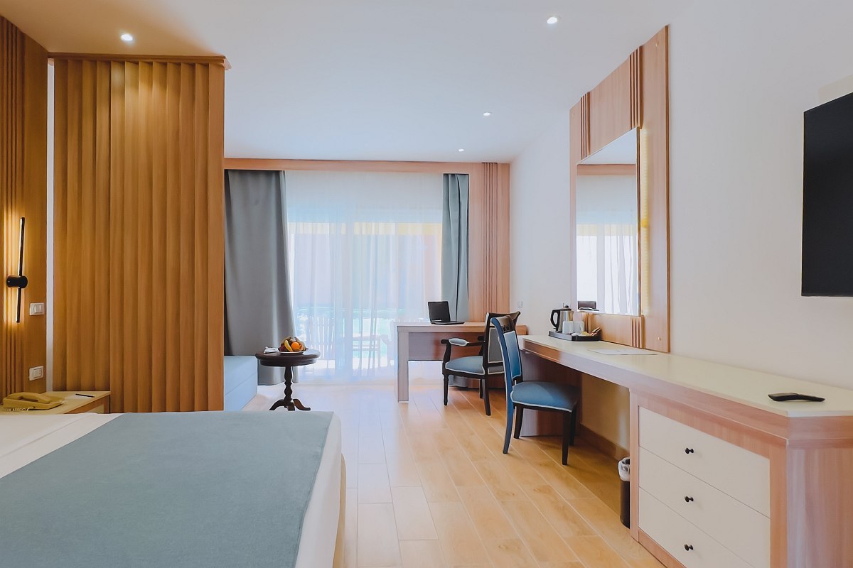 Bright, modern room with a large bed, light wood flooring, and a spacious layout. Features a desk with chairs, a TV, and a balcony with sheer curtains letting in natural light. Clean and airy design.