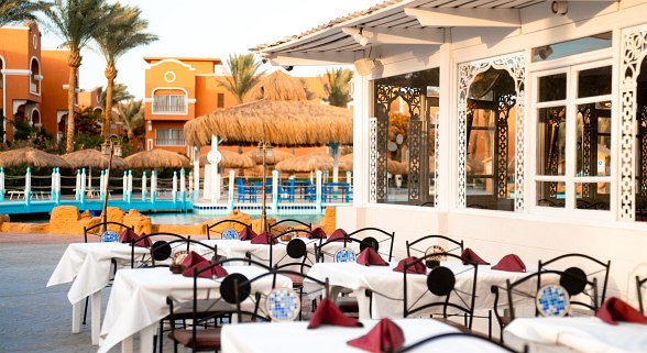 Outdoor dining area with white tablecloths and red napkins on black wrought iron chairs. Tropical setting with straw umbrellas and pool in the background.