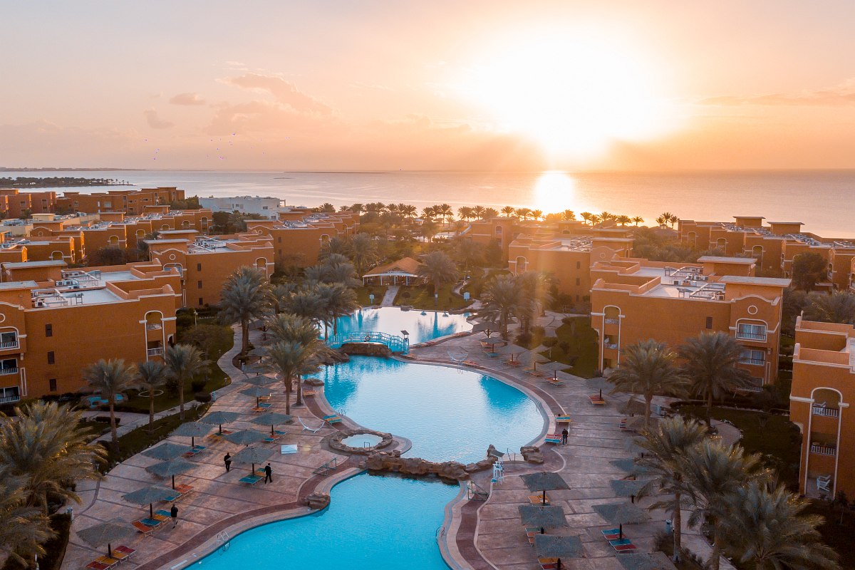 Sunset view of a resort with large pools, surrounded by orange buildings and palm trees, overlooking the sea.