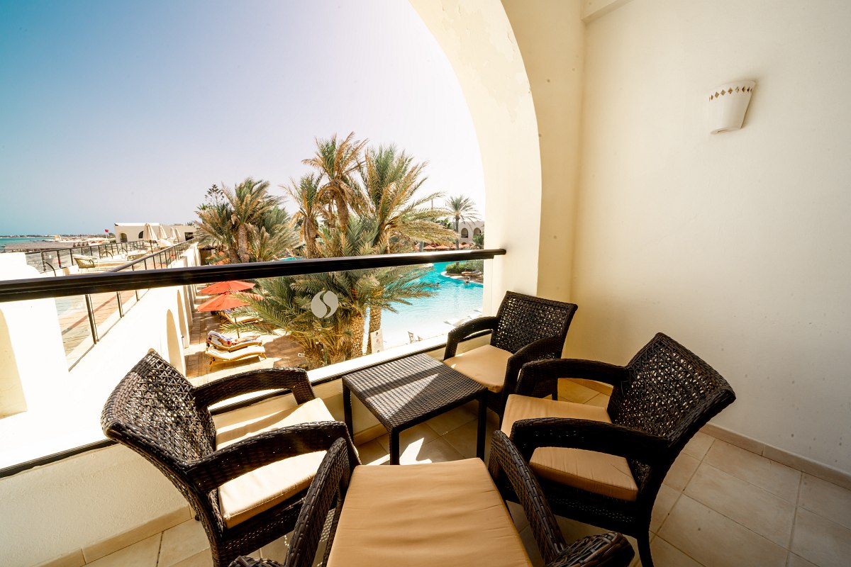 Balcony with seating area and view of palm trees, pool and sea in a sunny hotel complex.