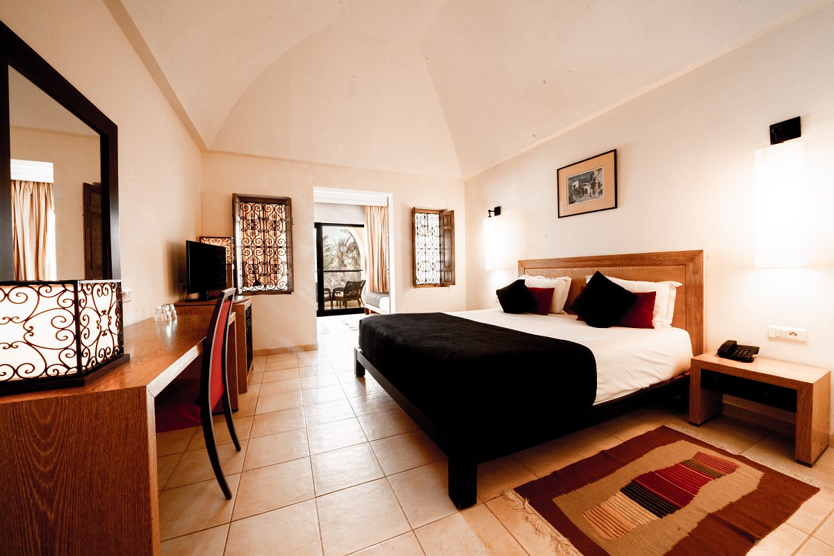 Hotel room with double bed, desk, carpet and simple furnishings in warm natural tones.