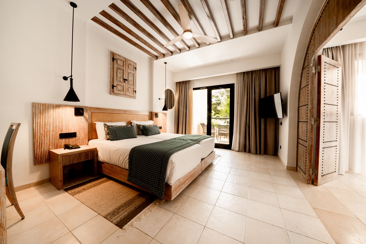 Bright hotel room with double bed, wooden elements, ceiling fan and balcony with garden view.