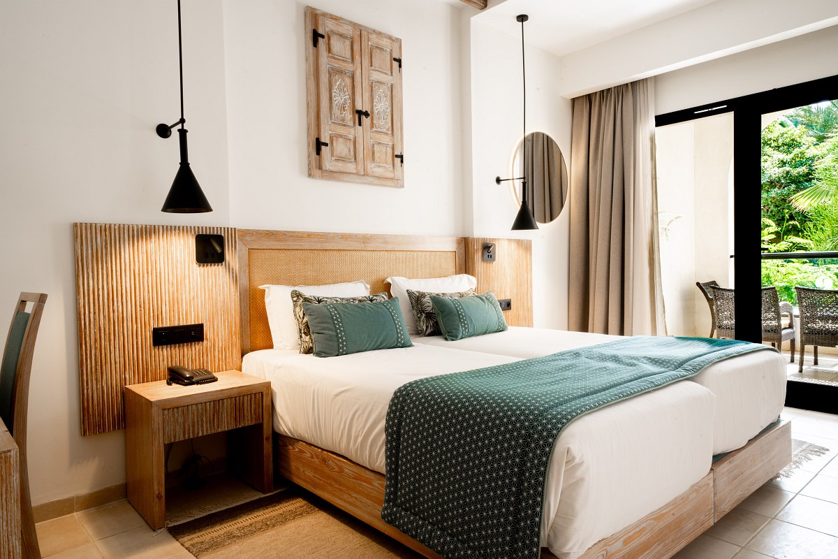Bright hotel room with double bed, wooden elements, ceiling fan and balcony with garden view.