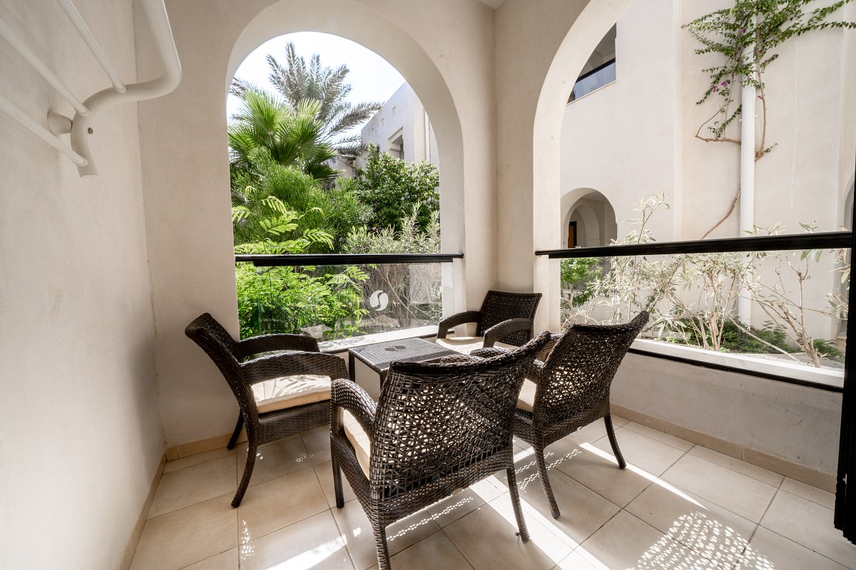 Covered hotel balcony with seating area and view of the green inner courtyard with palm trees and plants.