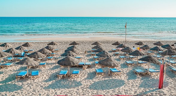 Sandy beach with parasols, sun loungers and beach volleyball court in front of a turquoise blue sea and clear sky.