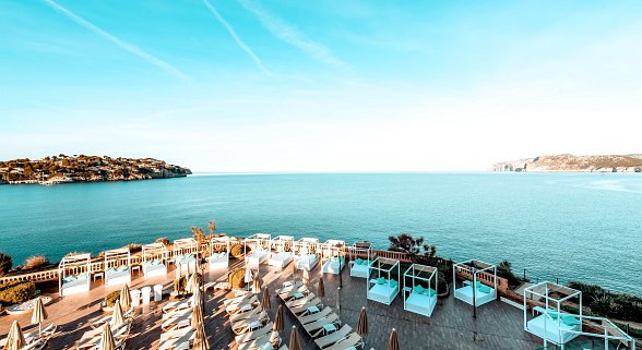 Sun terrace of a hotel with loungers, daybeds and sea view, surrounded by turquoise blue water and coastal landscape.