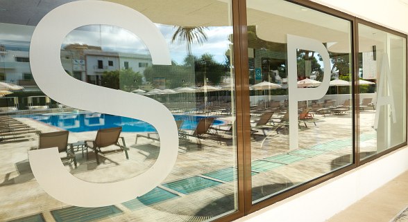 Large window front with “SPA” lettering, pool area with sun loungers and parasols behind it, palm trees in the background.
