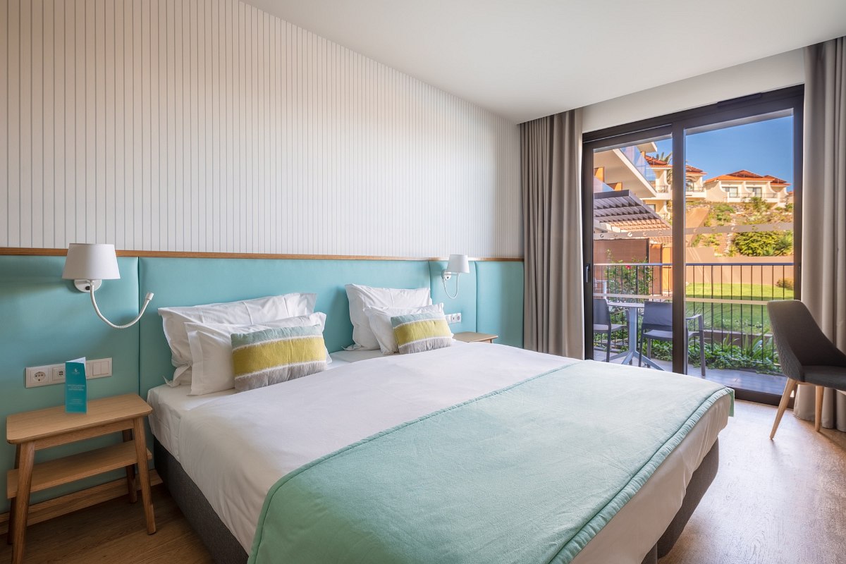 Modern hotel room with fresh turquoise and beige tones, large bed, terrace with garden view and plenty of natural light.