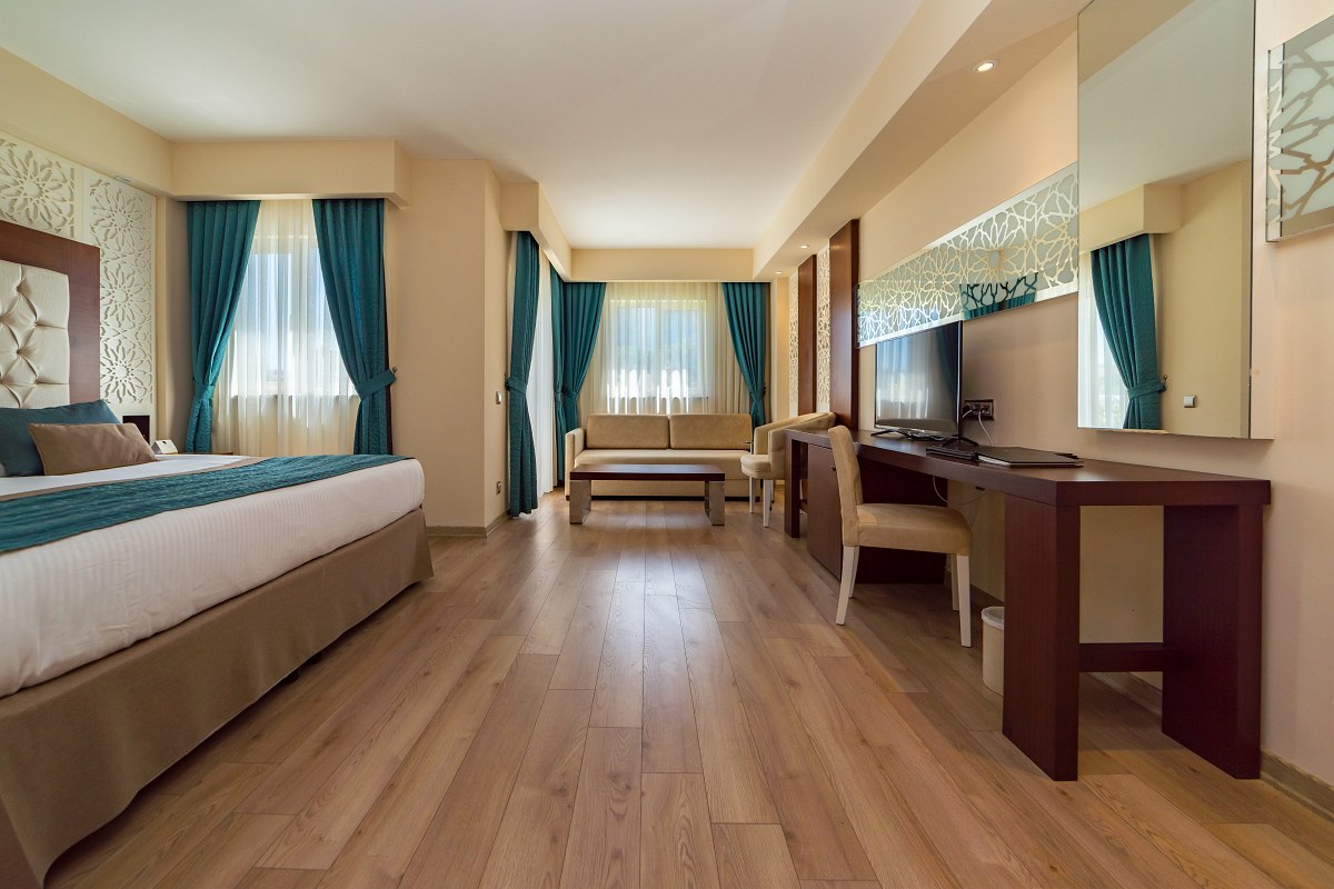 Bright, spacious hotel room with double bed, sofa, desk, TV and turquoise curtains.