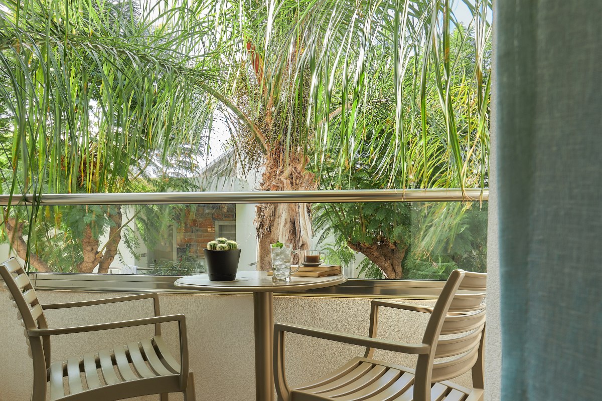Small balcony with two chairs and table, surrounded by palm trees and green trees with a view of neighboring buildings.