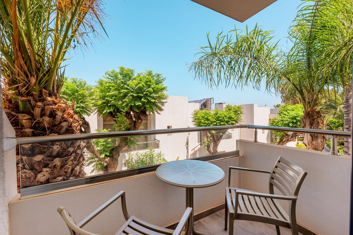 Small balcony with two chairs and table, surrounded by palm trees and green trees with a view of neighboring buildings.