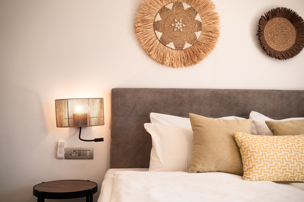 Close-up of a cozy bed with grey headboard, beige cushions, wall decoration made of natural material and reading lamp.