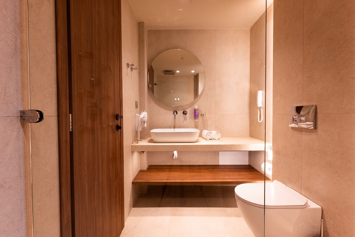 Modern hotel bathroom with round mirror light, white washbasin, wooden details and large walk-in shower.
