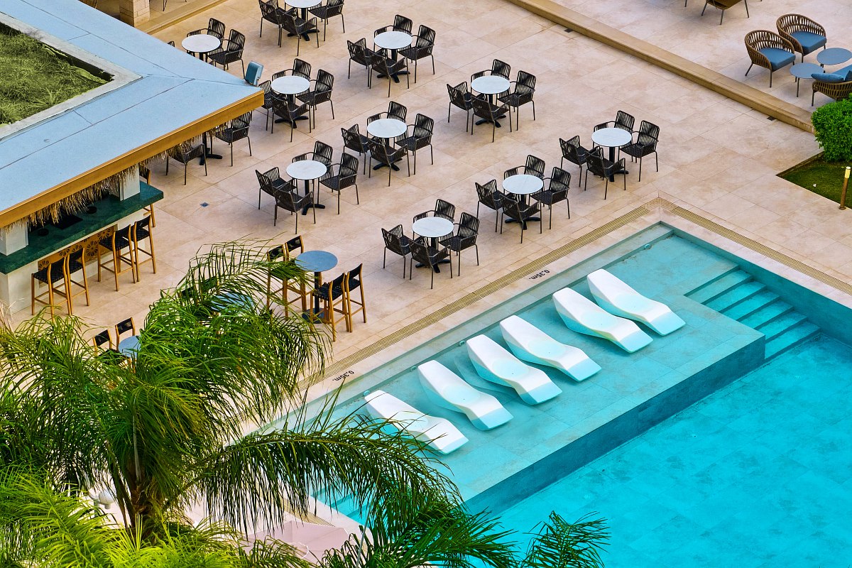 Large pool with sun loungers, bar and seating areas under parasols, surrounded by palm trees and Mediterranean buildings.