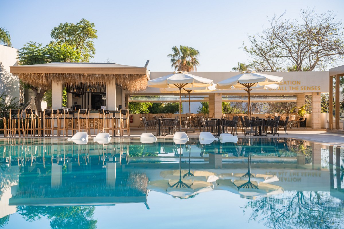 Large pool with sun loungers, bar and seating areas under parasols, surrounded by palm trees and Mediterranean buildings.