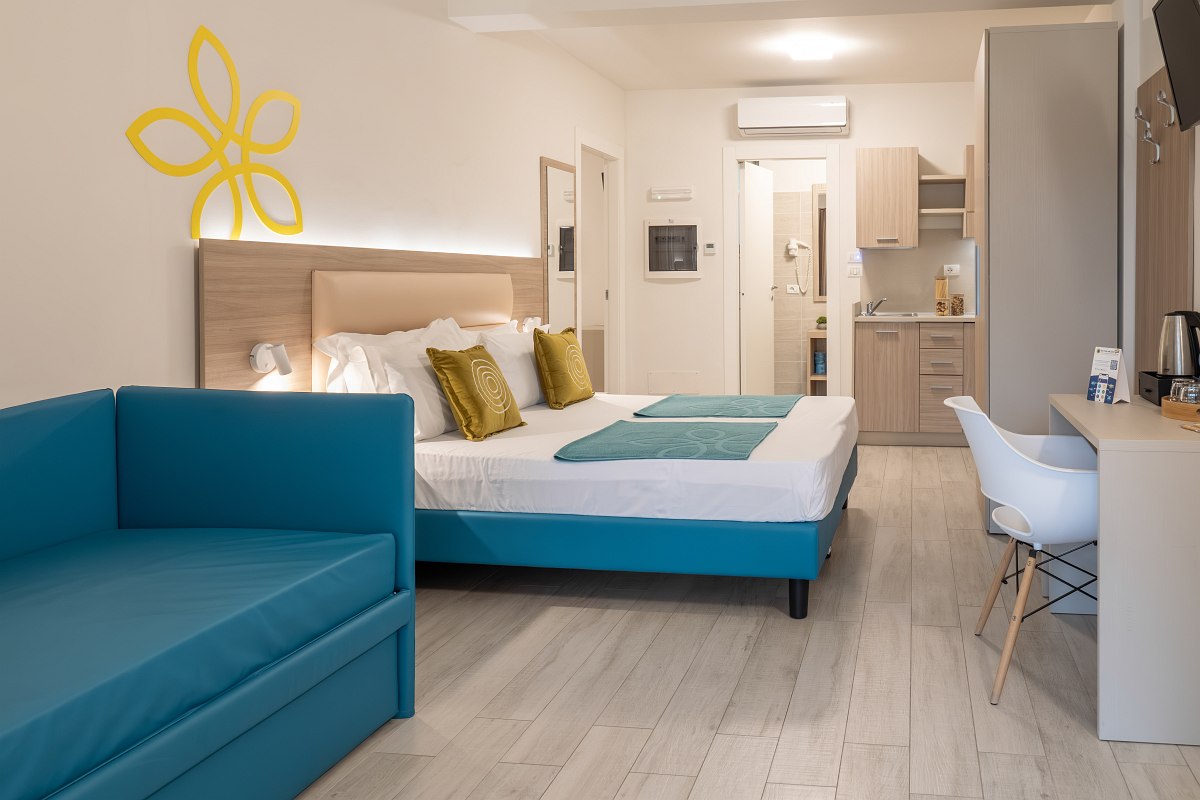 Modern furnished studio with a large double bed, turquoise sofa, desk with chair and a small kitchenette. The room is large, bright & friendly.