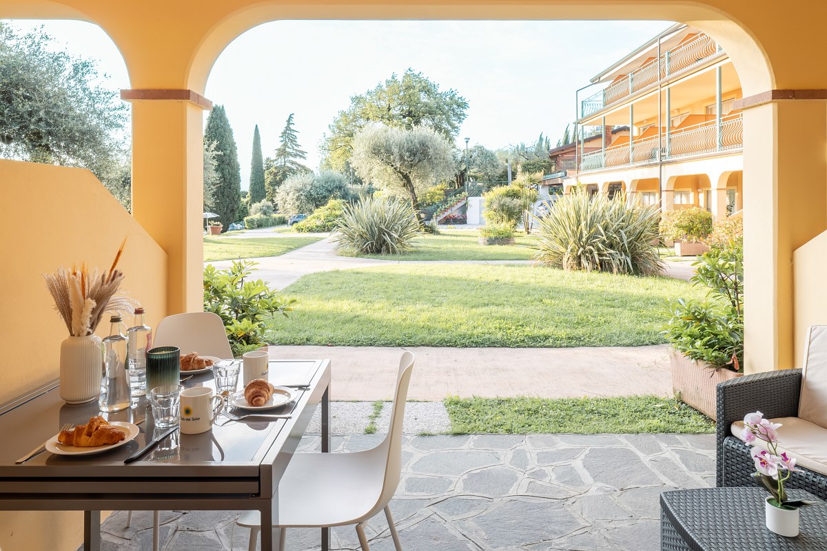 Covered and furnished terrace overlooking a well-tended garden. Table with chairs and lounge. Access to the lawn is at ground level and surrounded by wide paths.