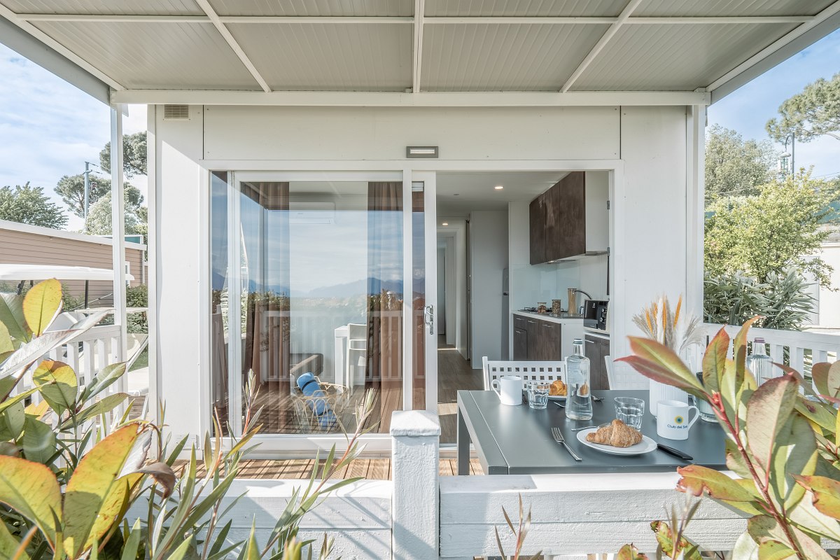 Covered terrace of a mobile home with breakfast table, view inside with kitchen and living area through glass front.