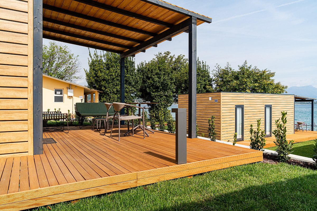 Modern wooden terrace with covered dining area, surrounded by lawn and views of other mobile homes and the lake.