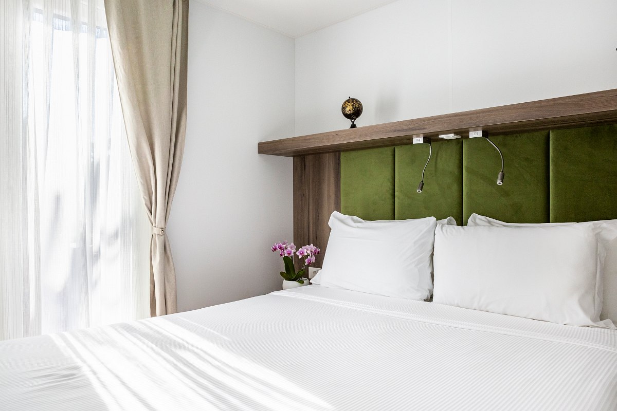 Bright bedroom with double bed, green upholstered headboard, shelf with reading lamps and daylight through curtained window.