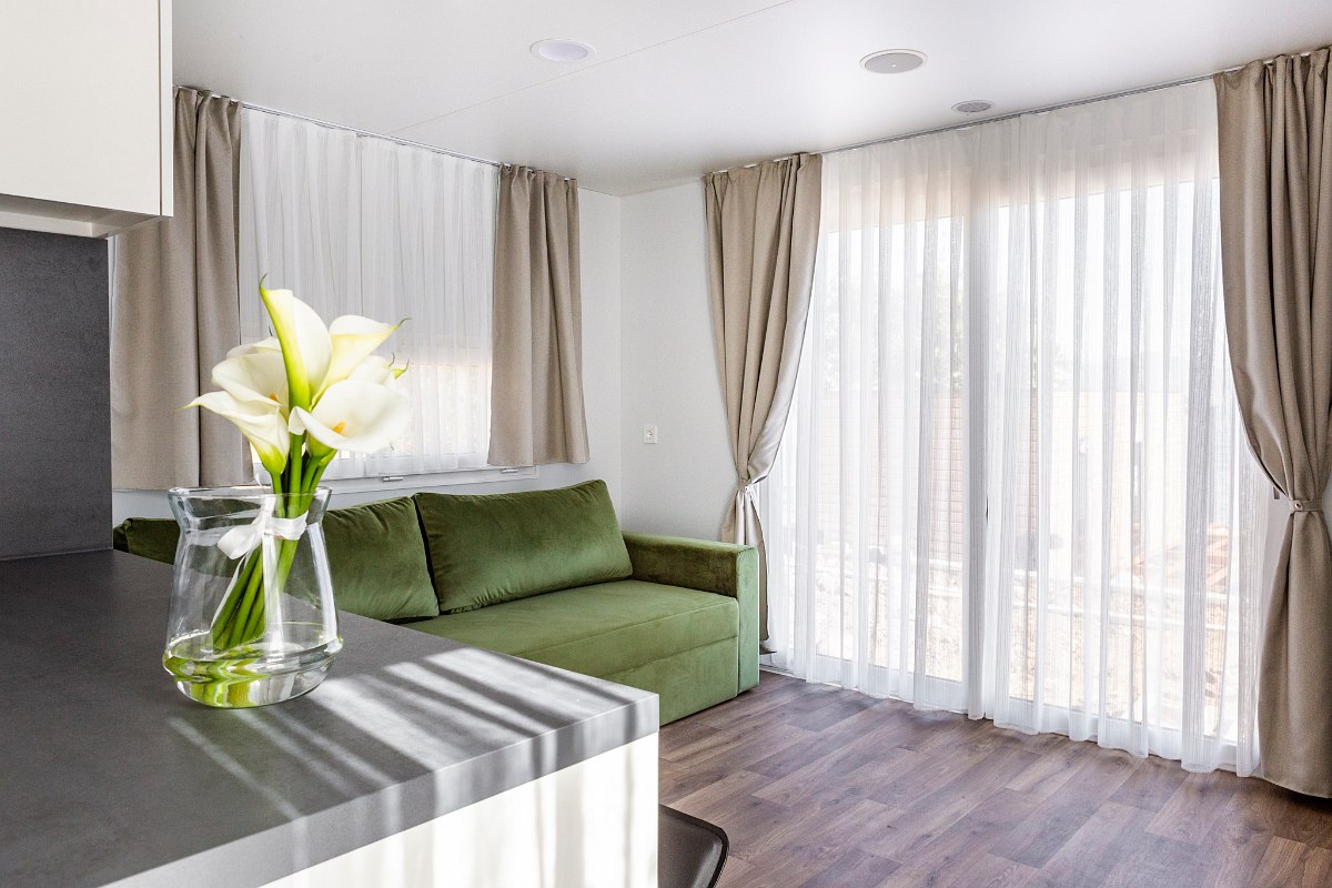 Bright living area with green sofa, floor-to-ceiling windows with curtains and flowers on the kitchen worktop.