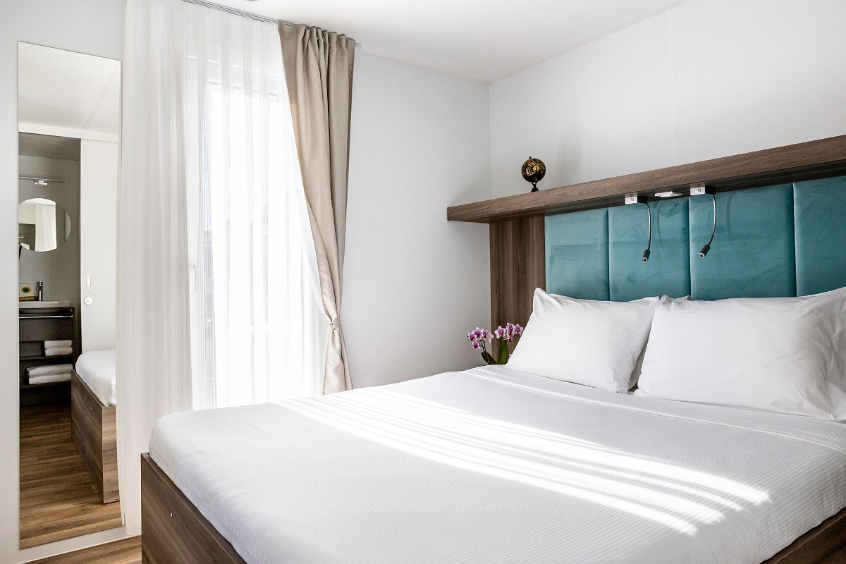 Bright bedroom with double bed, turquoise headboard, mirror, natural light and view of the adjoining bathroom.