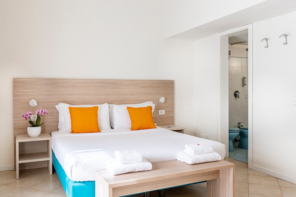 Bright hotel room with double bed, orange pillows, flowers and direct access to a modern bathroom.