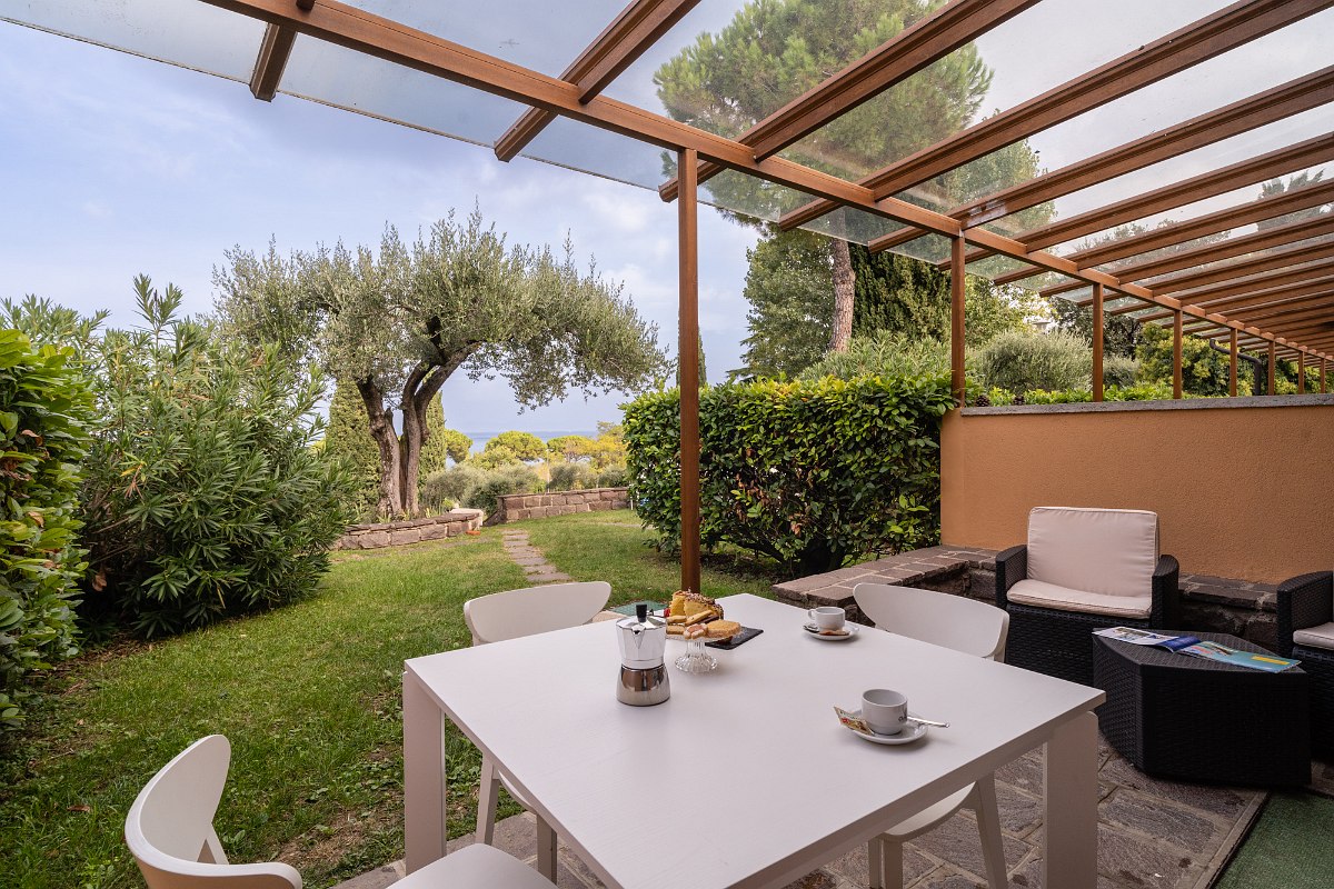 Covered terrace with seating area, breakfast table and view of the green garden with olive tree.