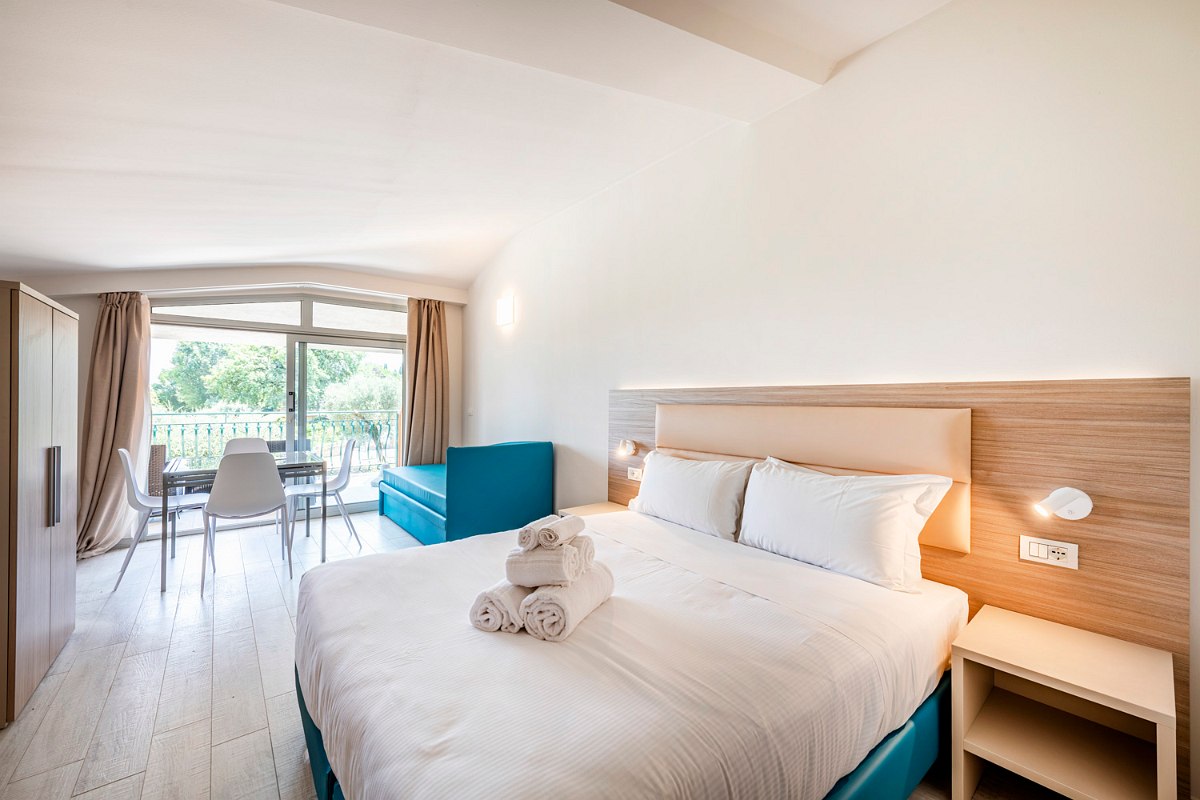 Modern studio with double bed, turquoise sofa, large mirror, bedside tables, air conditioning, TV and desk and lake view.