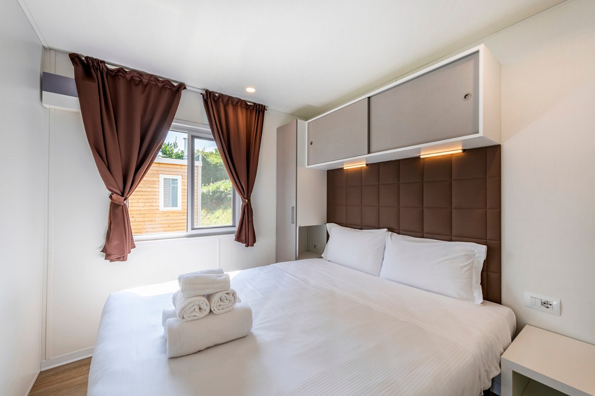 Compact bedroom with double bed, window, brown curtains and storage space above the bed - bright and modern.