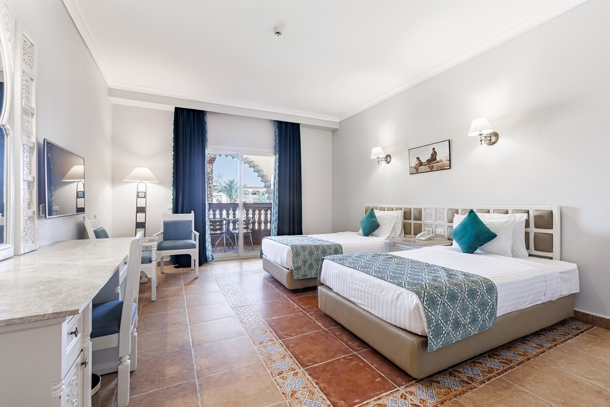 A charming hotel room with two single beds, patterned bedspreads, wall lamps, a TV, a vanity with a mirror, and a small table and chairs by sliding glass doors opening to a balcony.