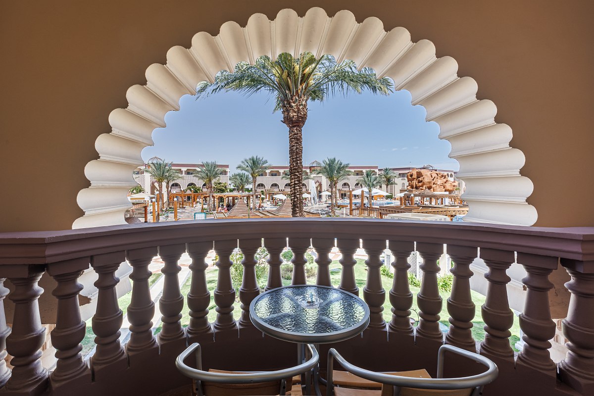 A charming hotel room with two single beds, patterned bedspreads, wall lamps, a TV, a vanity with a mirror, and a small table and chairs by sliding glass doors opening to a balcony.