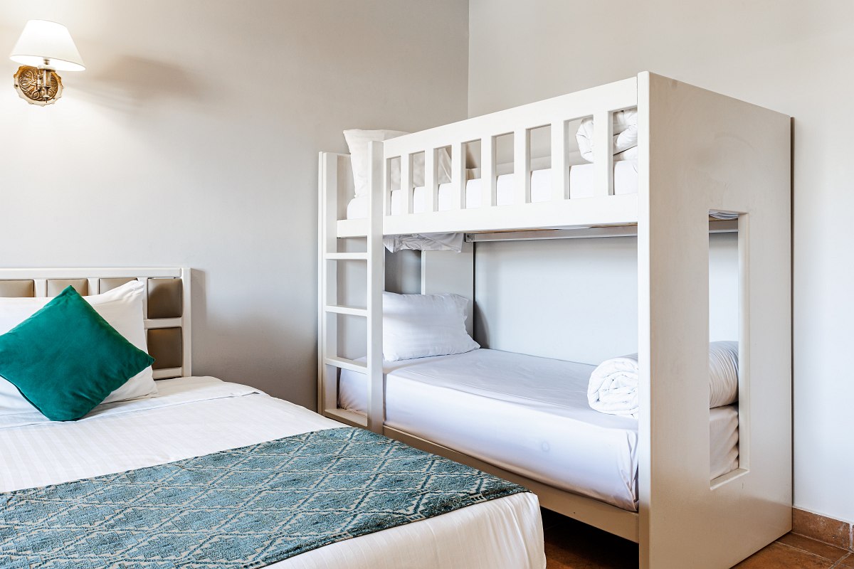 A cozy balcony with two striped lounge chairs and a small glass table, overlooking a sunny beach filled with white umbrellas and loungers beside the blue sea.