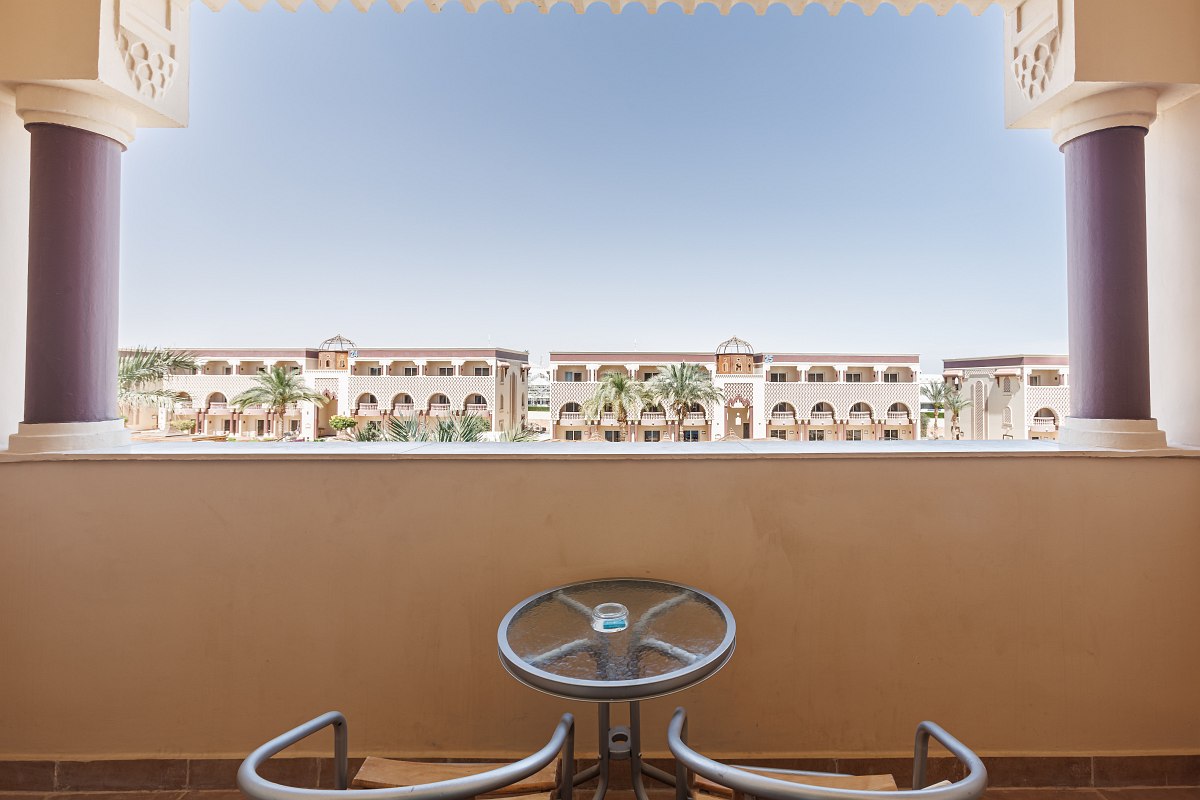 A cozy balcony with two striped lounge chairs and a small glass table, overlooking a sunny beach filled with white umbrellas and loungers beside the blue sea.