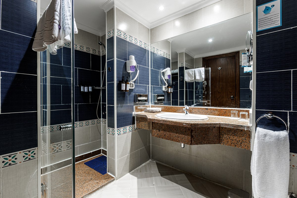 A bright, cozy hotel room with a large bed covered in white linens and a patterned throw, a TV on a stand, two cushioned armchairs, a floor lamp, and a small table with flowers, all set on a tiled floor with a colorful rug.