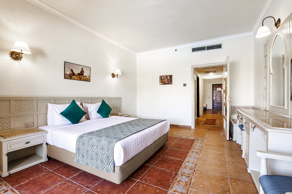 A charming balcony with a round glass table and two wicker chairs overlooks a lush garden and elegant hotel building under a clear sky. The balcony features a beautifully scalloped arch and ornate balustrades.