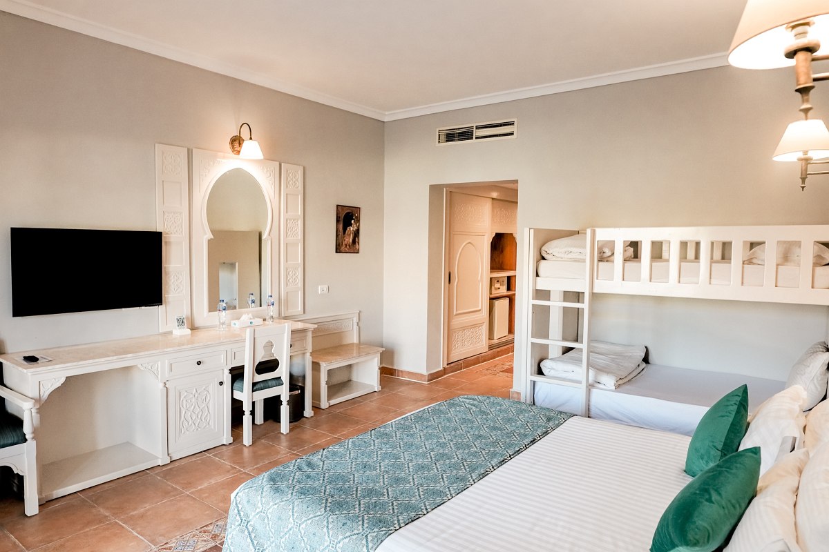 A warm, cozy room featuring two double beds and a bunk bed. The decor is classic with cream-colored furniture and tiled flooring, creating a welcoming atmosphere.