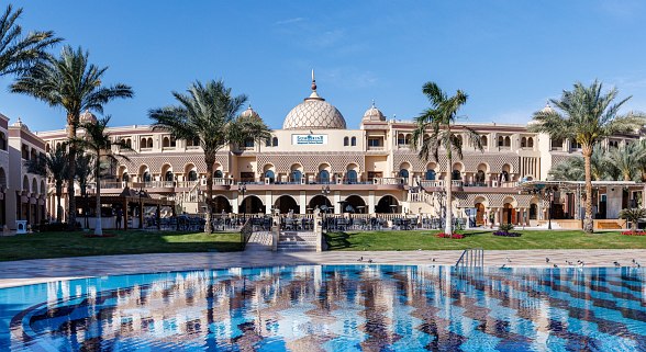 A stunning resort with a large central pool surrounded by palm trees, pergolas, and elegant buildings. The spacious outdoor seating area features a beautiful star-shaped fountain, perfect for relaxation and socializing.