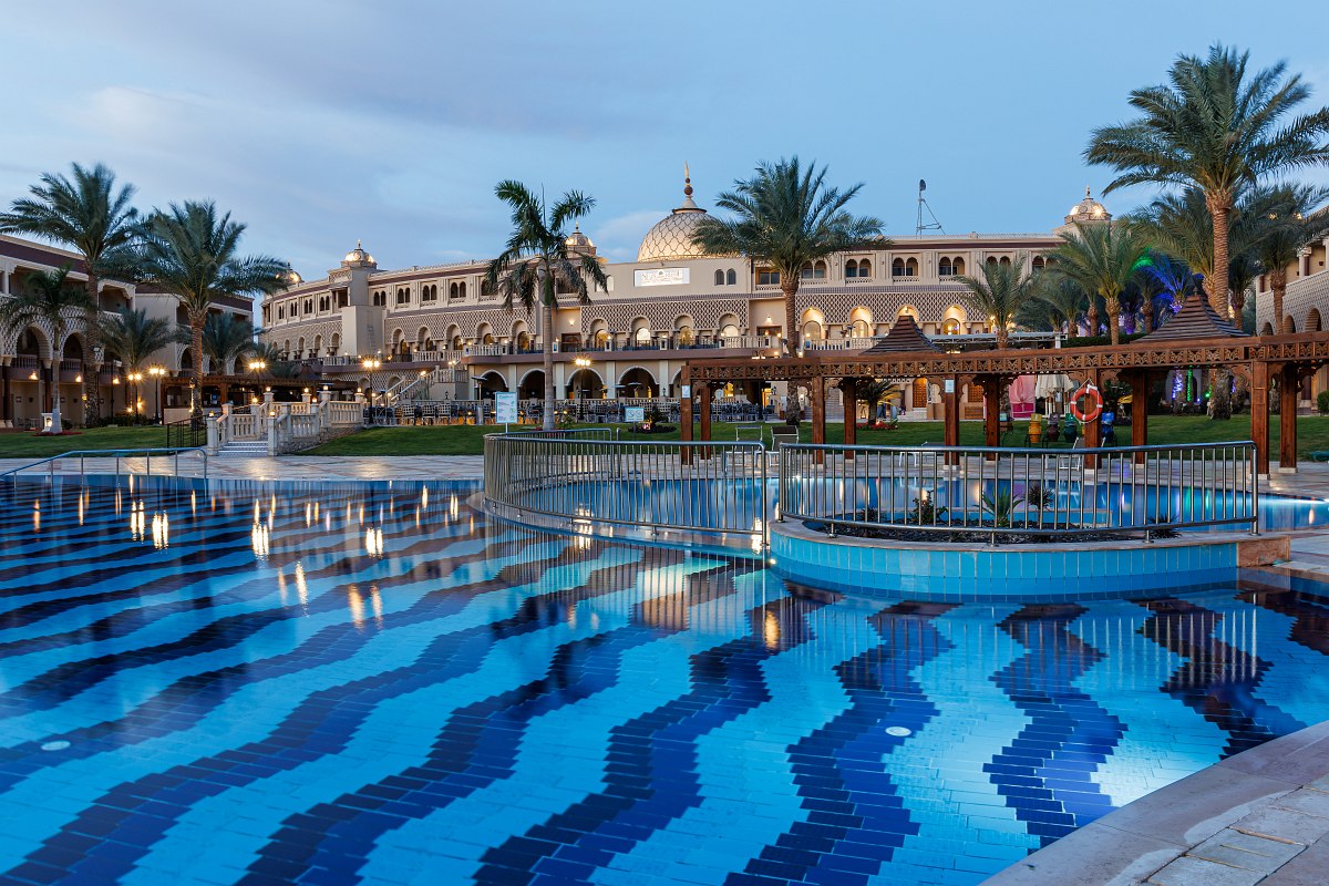 Large resort with beautiful pool, palm trees, sun loungers, and shaded seating areas in a peaceful, sunny setting.