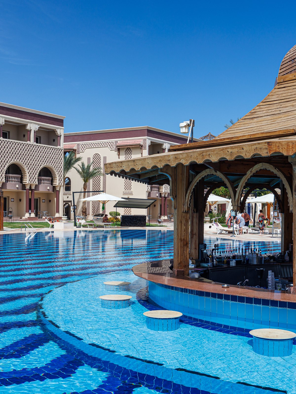 Pool bar with submerged stools and wooden canopy, surrounded by sun loungers and elegant resort architecture reflected in the clear blue water.